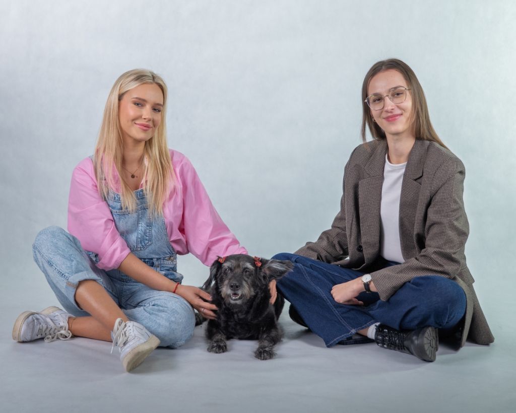 Na zdjęciu widoczne są dwie osoby siedzące na podłodze na jednolitym, jasnym tle. Między nimi znajduje się mały czarny pies. Obie osoby mają swobodną, nieformalną postawę i delikatnie głaszczą zwierzę. Scena ma spokojny, przyjazny charakter.